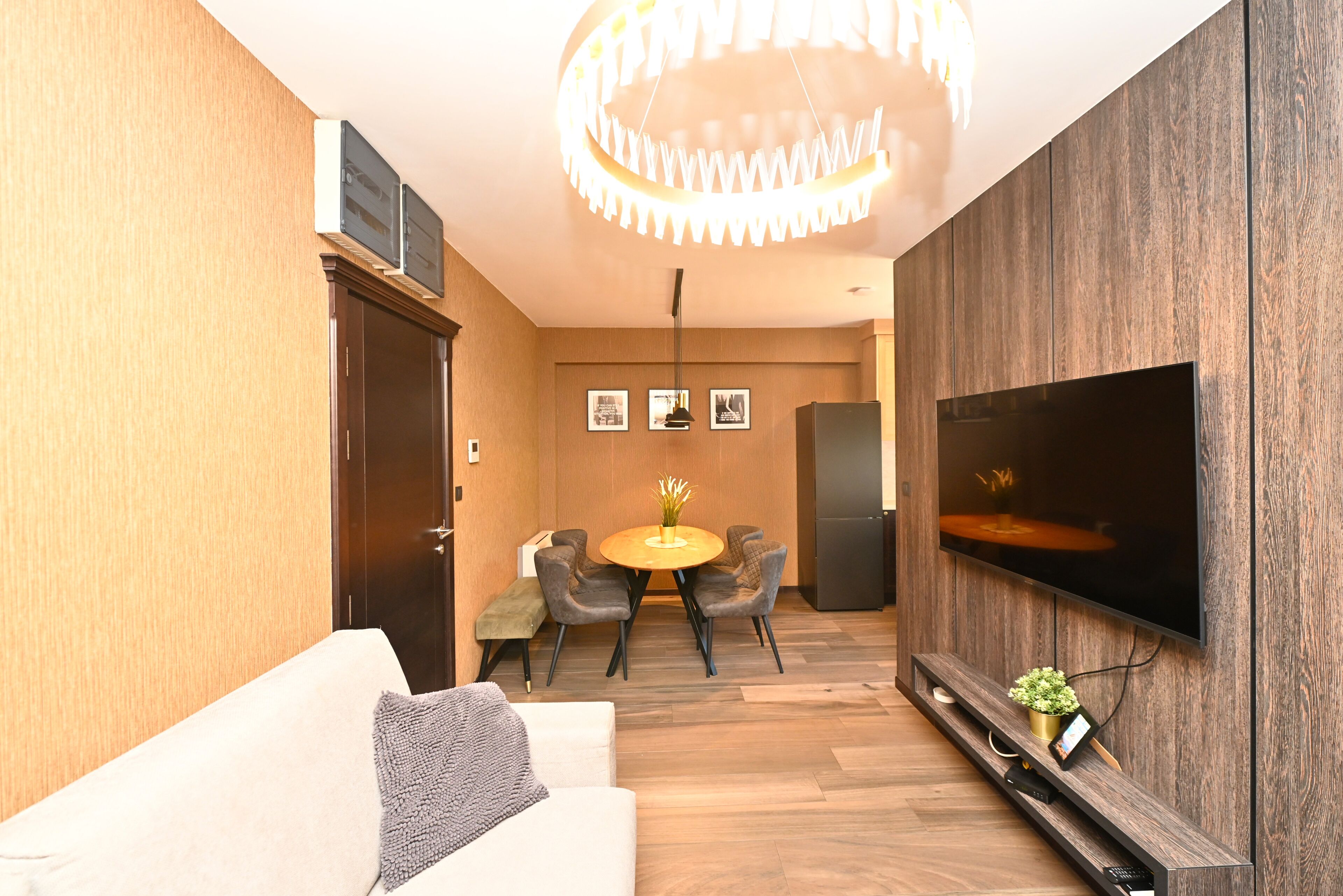 Uma sala de estar moderna com um sofá, uma mesa de jantar com cadeiras, uma TV fixada na parede e uma luminária decorativa no teto.
