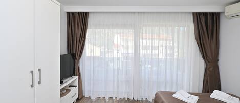 Quarto de hotel com cama, televisão em suporte, guarda-roupa e janela com cortinas.