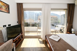 A hotel room with a TV, a bed, a balcony with a table and chairs, and a view of buildings and mountains.
