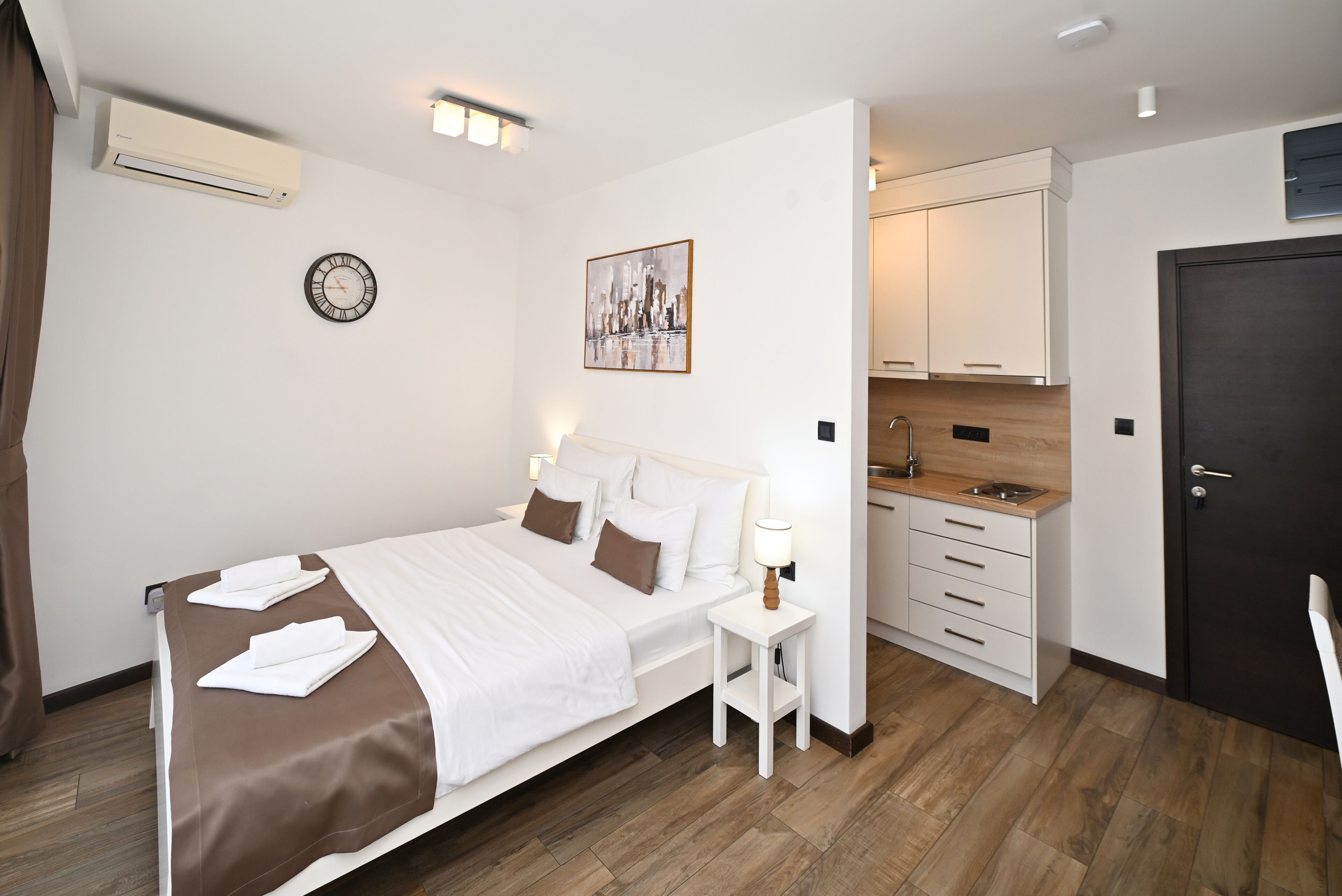 Habitación de hotel con cama, mesita de noche, reloj de pared, una obra de arte y una kitchenette.