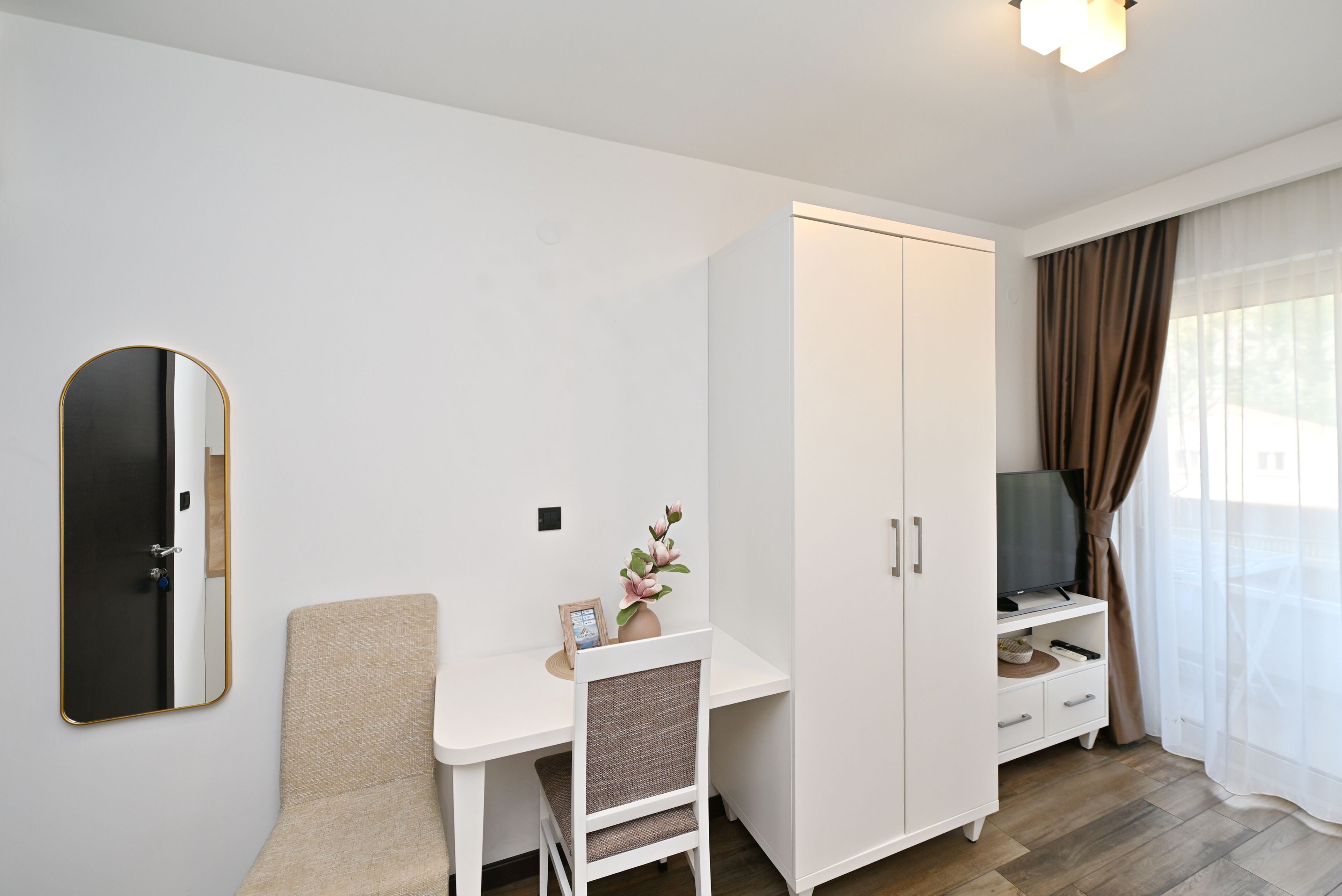 A room with a white desk, a chair, a wardrobe, a television, and a window with curtains.