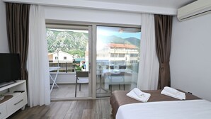 A hotel room with a bed, a TV, a balcony with a table and chairs, and a view of buildings and mountains. - Casa Nuova (Kotor)