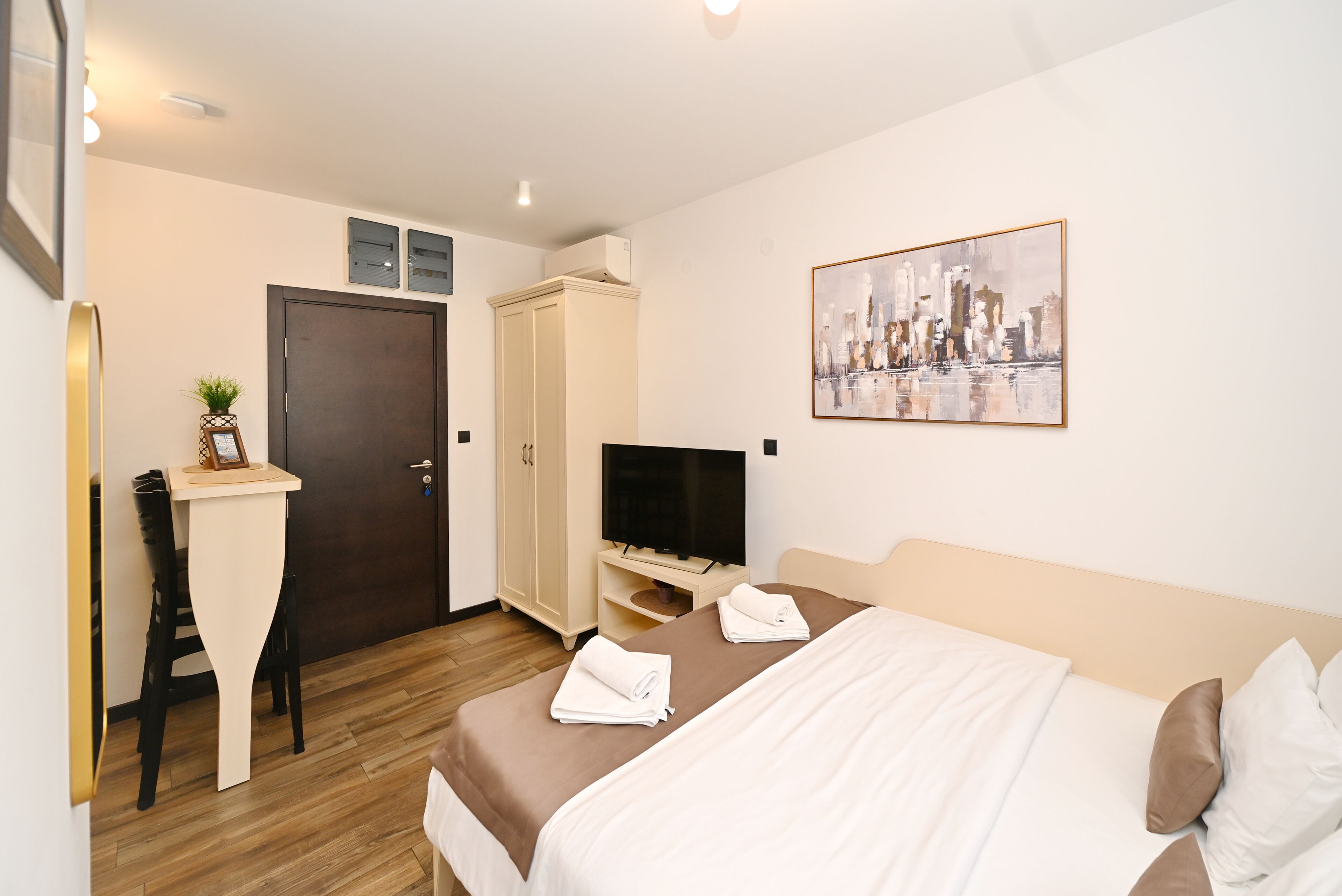 A hotel room with a bed, a TV, a desk with a chair, a wardrobe, and a painting on the wall.
