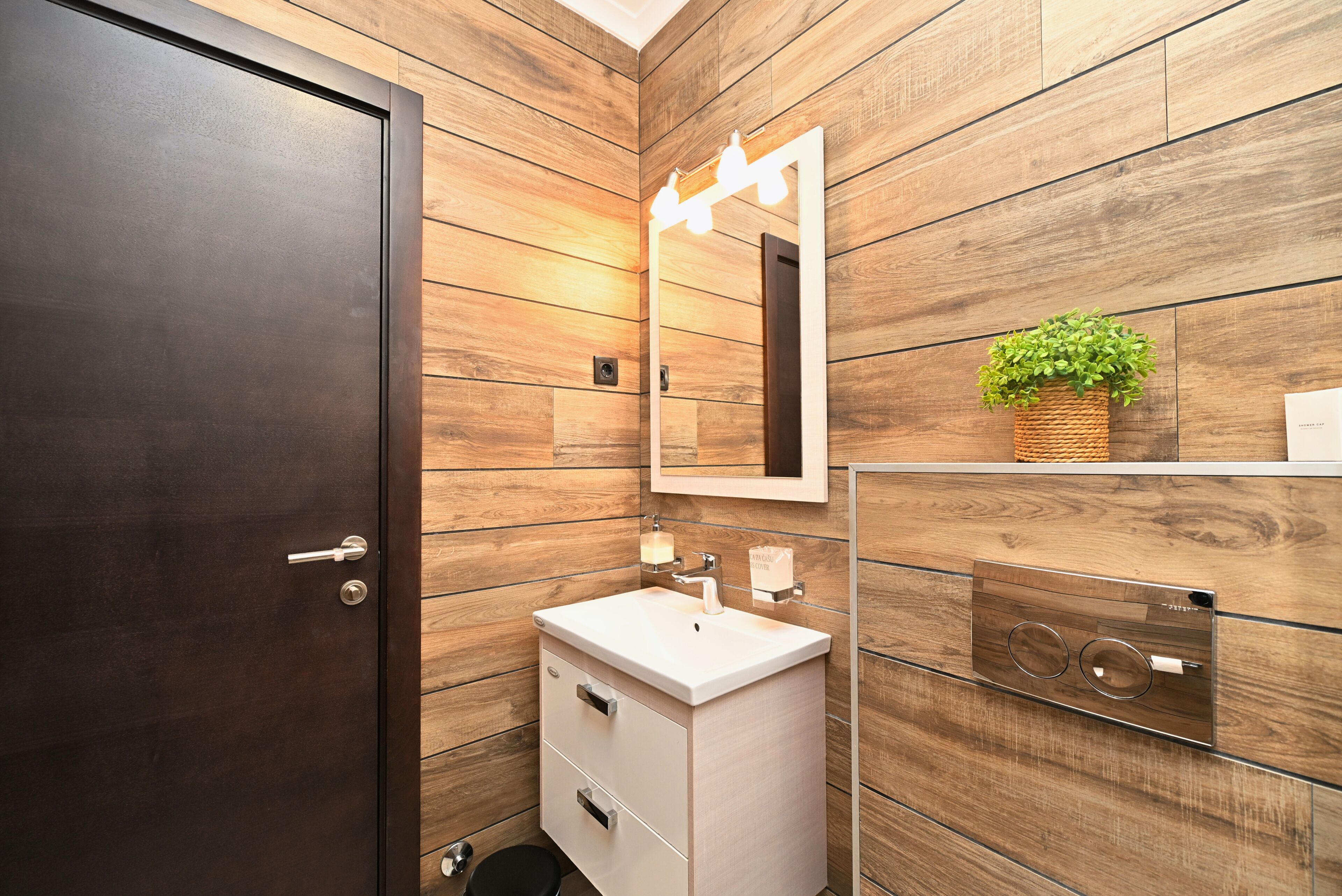 Un baño con paneles de madera, un lavamanos blanco, un espejo con luces, una planta pequeña y una puerta.