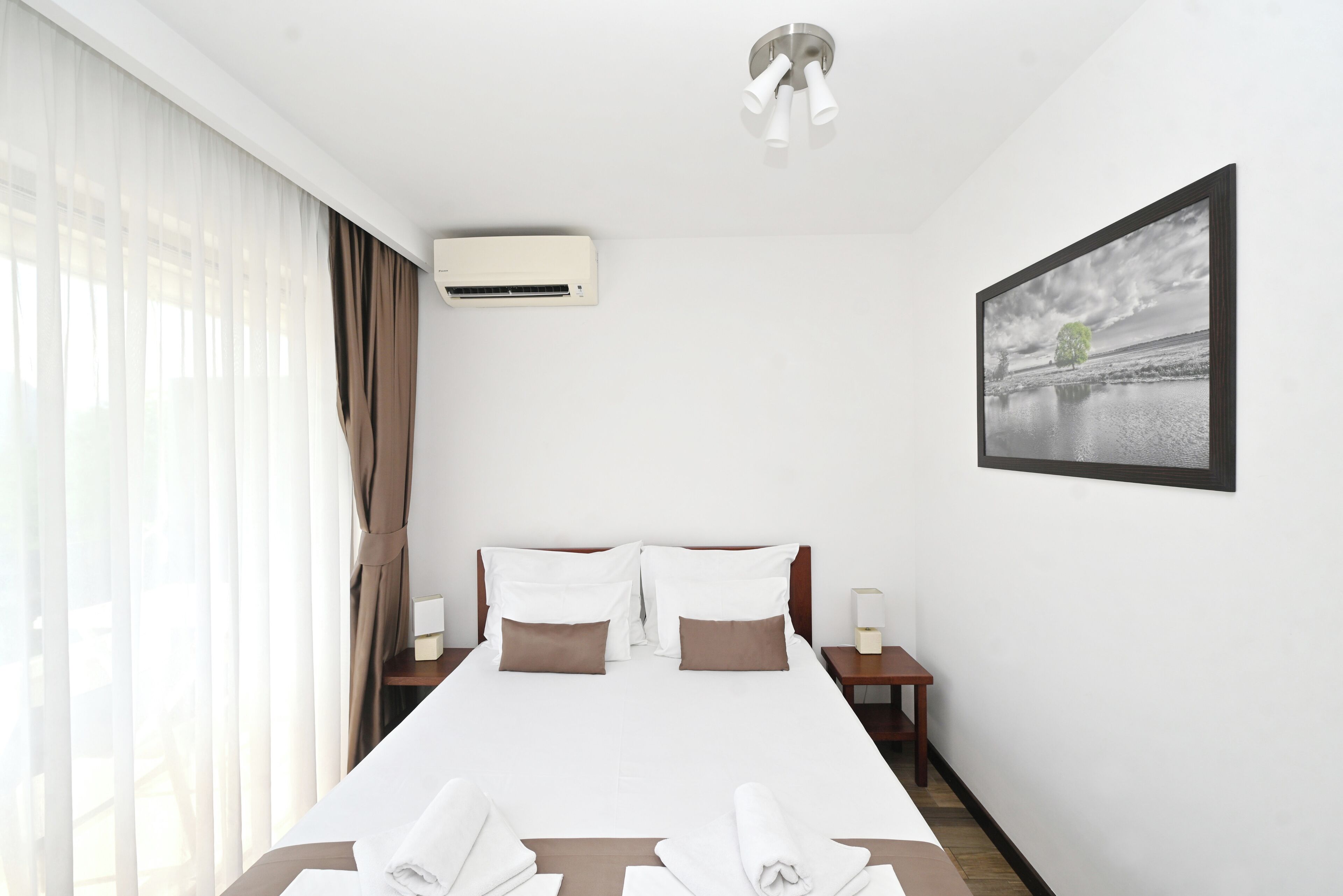 Habitación de hotel con cama, aire acondicionado, lámparas de noche, un cuadro enmarcado y una ventana con cortinas.