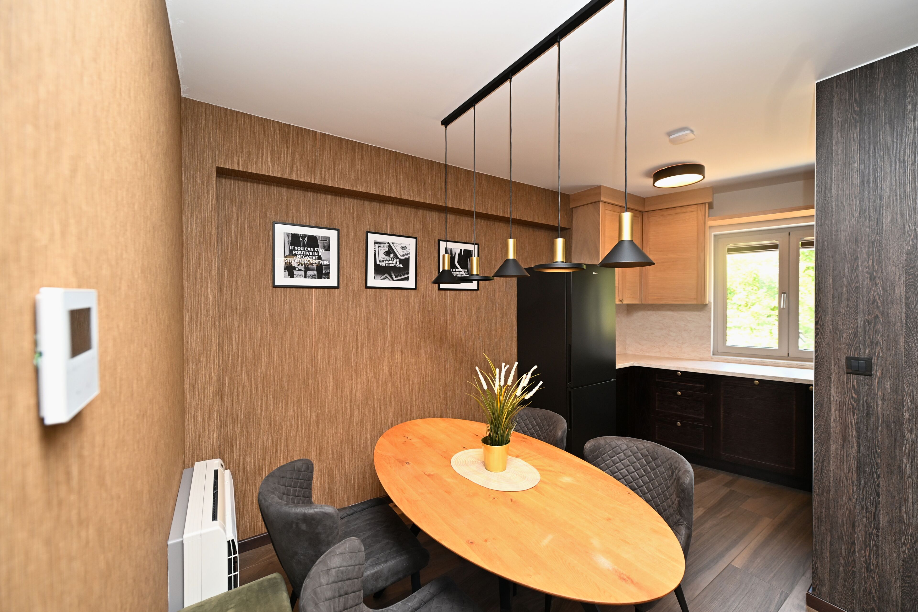 A modern kitchen with a wooden dining table, pendant lights, and framed pictures on the wall.