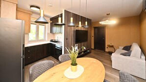 A modern kitchen with a built-in oven, a dining table with a vase of flowers, and a living area with a sofa and a TV. - Casa Nuova (Kotor)