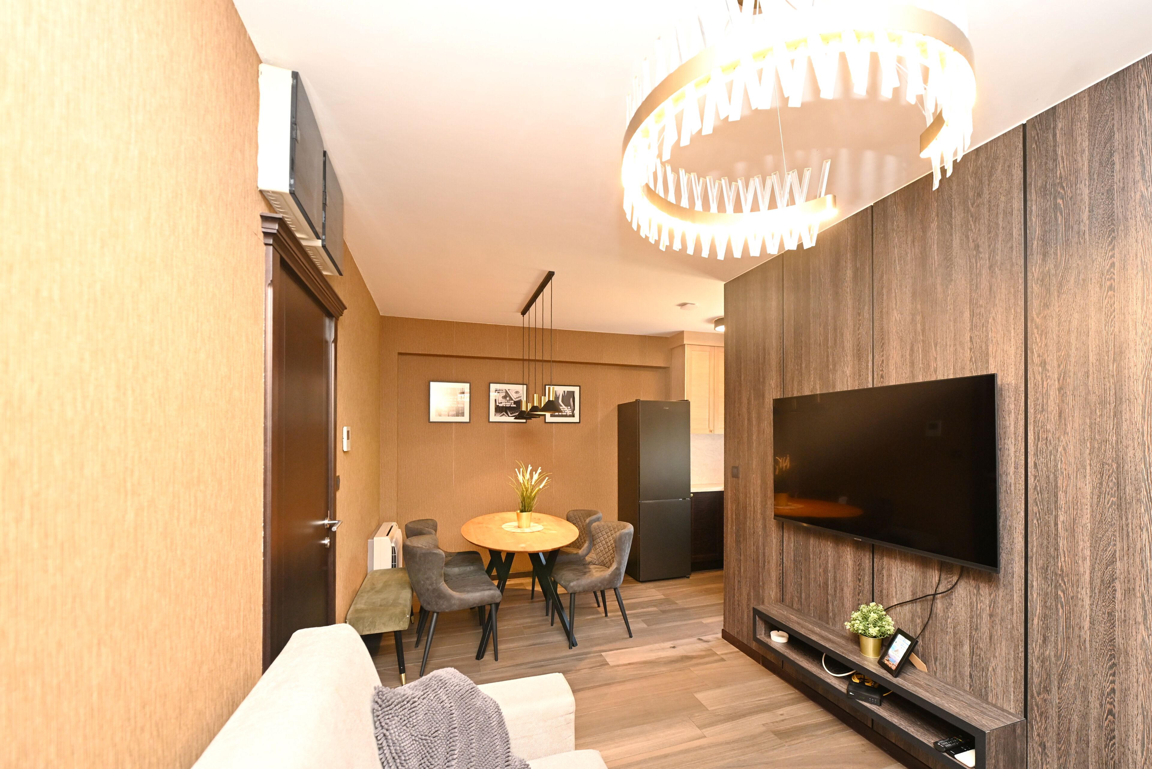 A modern living room with a flat-screen TV, wooden flooring, a round dining table, and a chandelier.