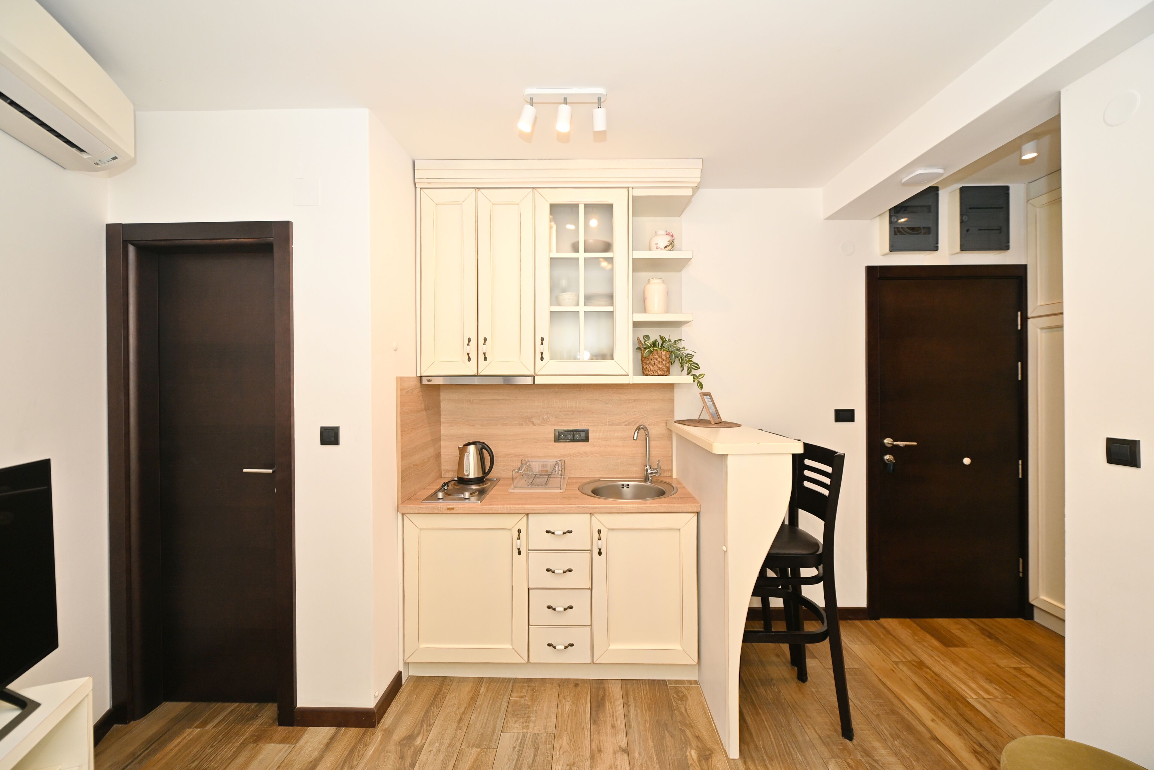 A compact kitchen with a wooden countertop, a sink, a stove, and a chair. There are cabinets with glass doors and a small shelf with decorative items.