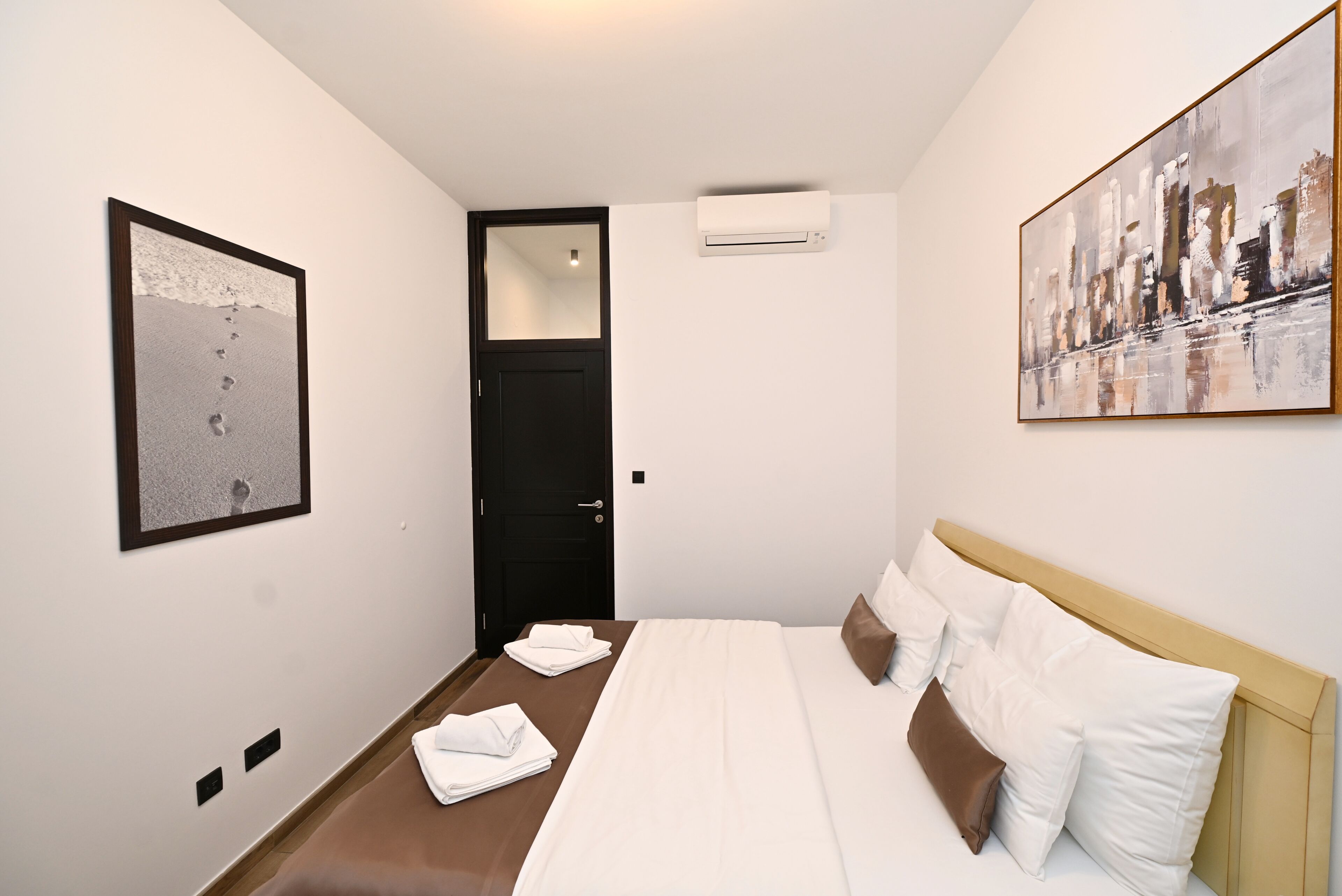 A hotel room with a bed, a door, an air conditioning unit, and two artworks on the walls.