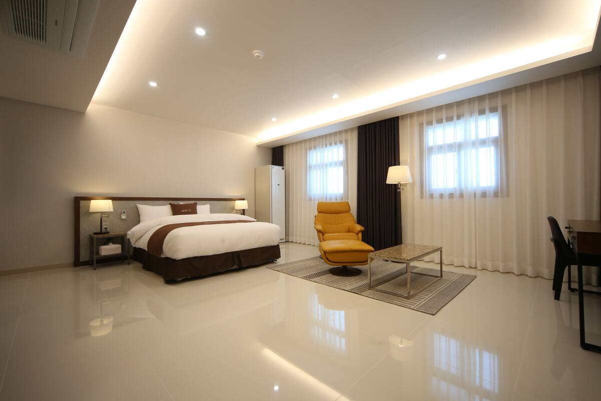 A modern hotel room with a large bed, a seating area with a chair and table, and a dining table with chairs.