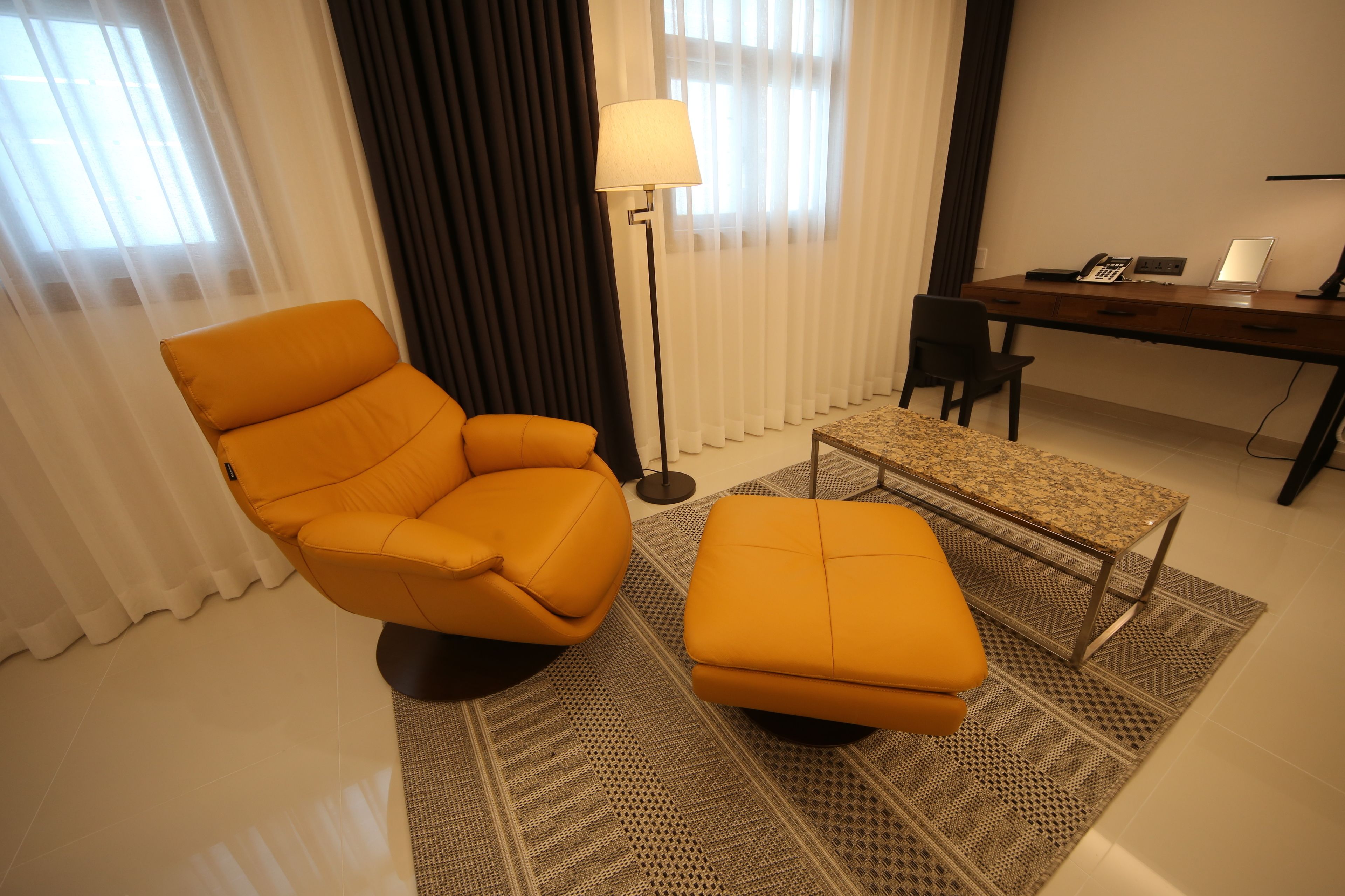 Een moderne woonkamer met een gele fauteuil, een passende voetenbankje, een vloerlamp, een bureau met een laptop en een kamerkleed met een patroon.