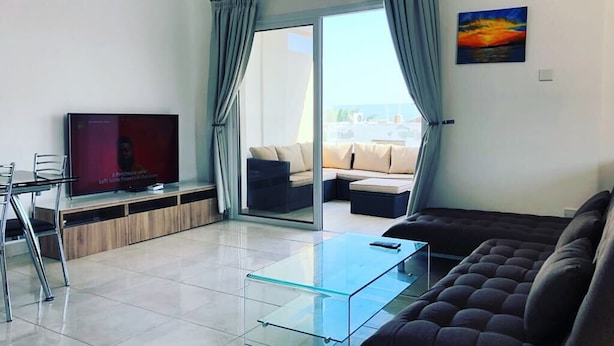 Una sala moderna con una mesa de centro de vidrio, un sofá oscuro, un televisor de pantalla plana y un ventanal corredero que da a un área de estar al aire libre.