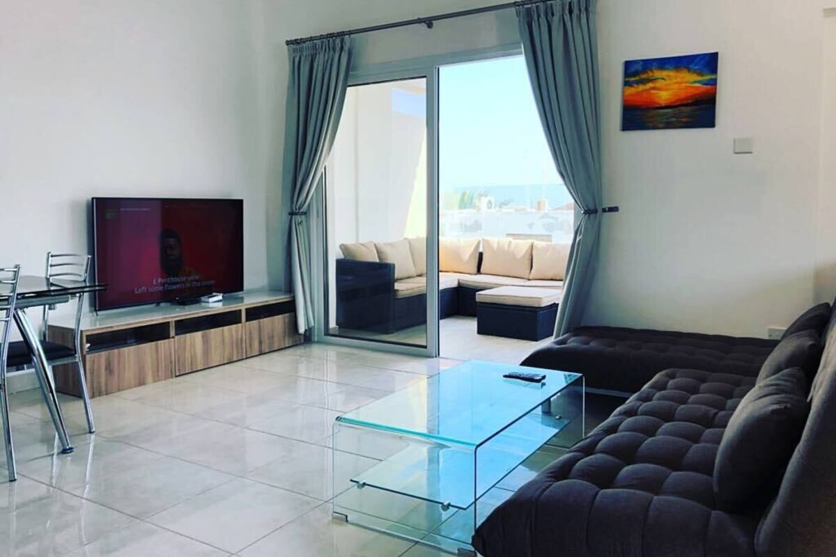 Un salon moderne avec une table basse en verre, un canapé foncé, un téléviseur à écran plat et une porte-fenêtre coulissante donnant sur un espace de détente extérieur.