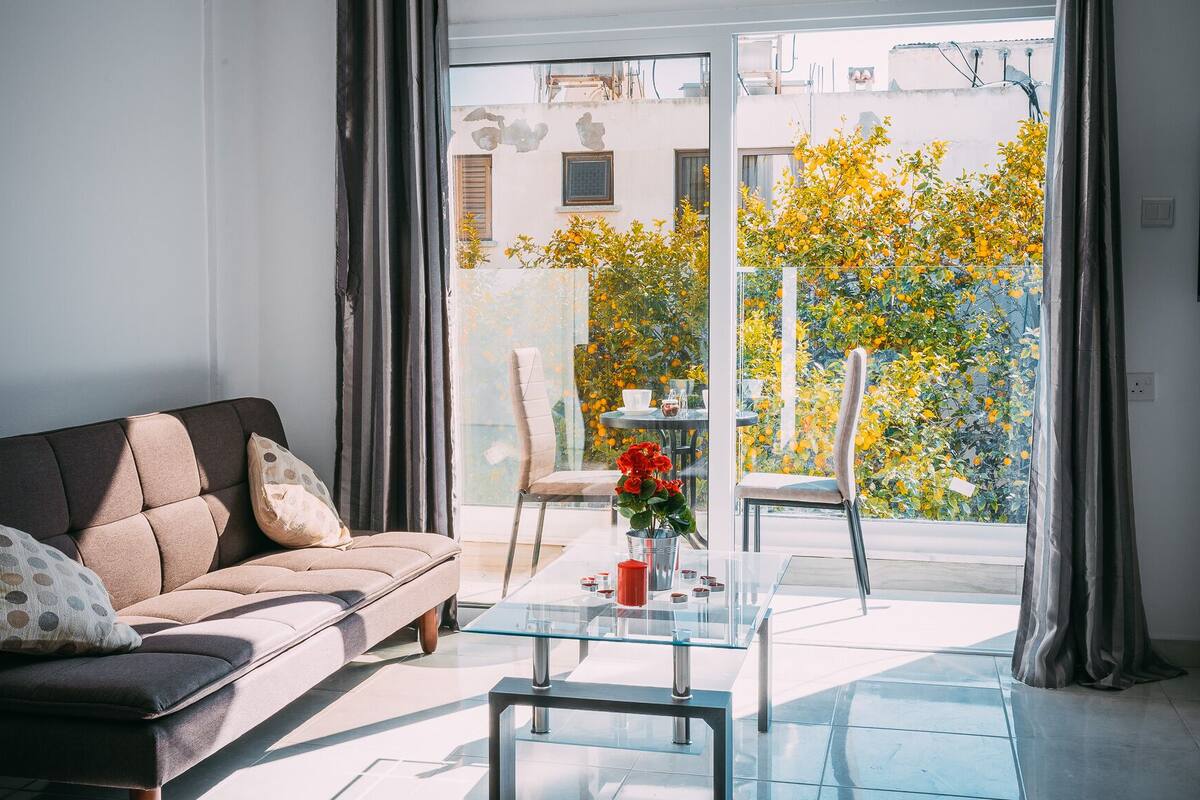Un salon moderne avec un canapé, une table basse en verre et une vue sur un jardin où poussent des fleurs jaunes.