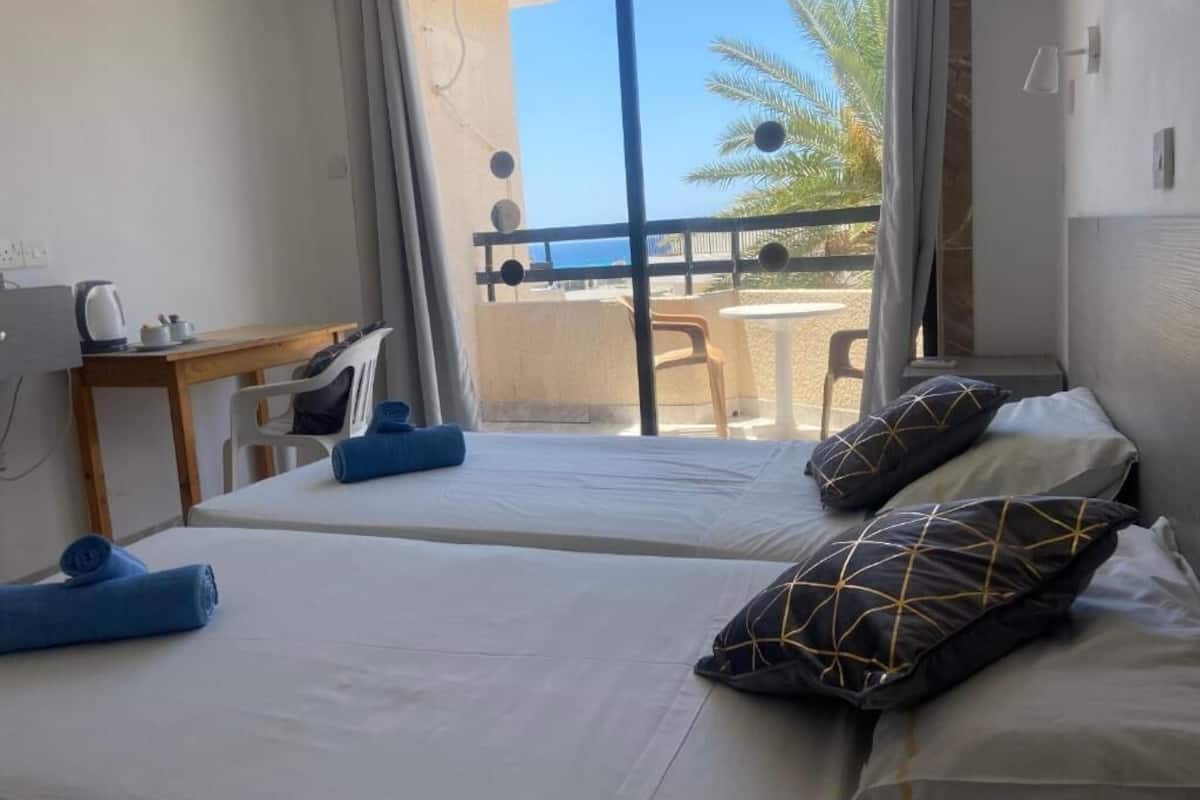 Une chambre avec deux lits, un bureau avec une chaise et une vue sur la mer depuis un balcon.