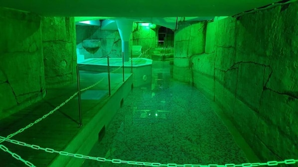 A dimly lit, narrow passageway with stone walls and a central water feature, enclosed by a chain barrier.
