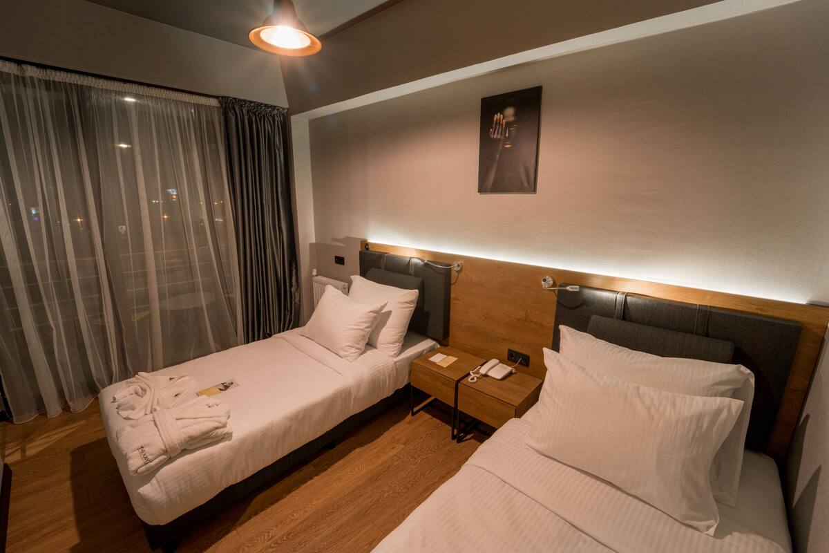 Ein Hotelzimmer mit zwei Betten, Holzboden, einem Nachttisch mit Telefon und einem Fenster mit durchsichtigen Vorhängen.
