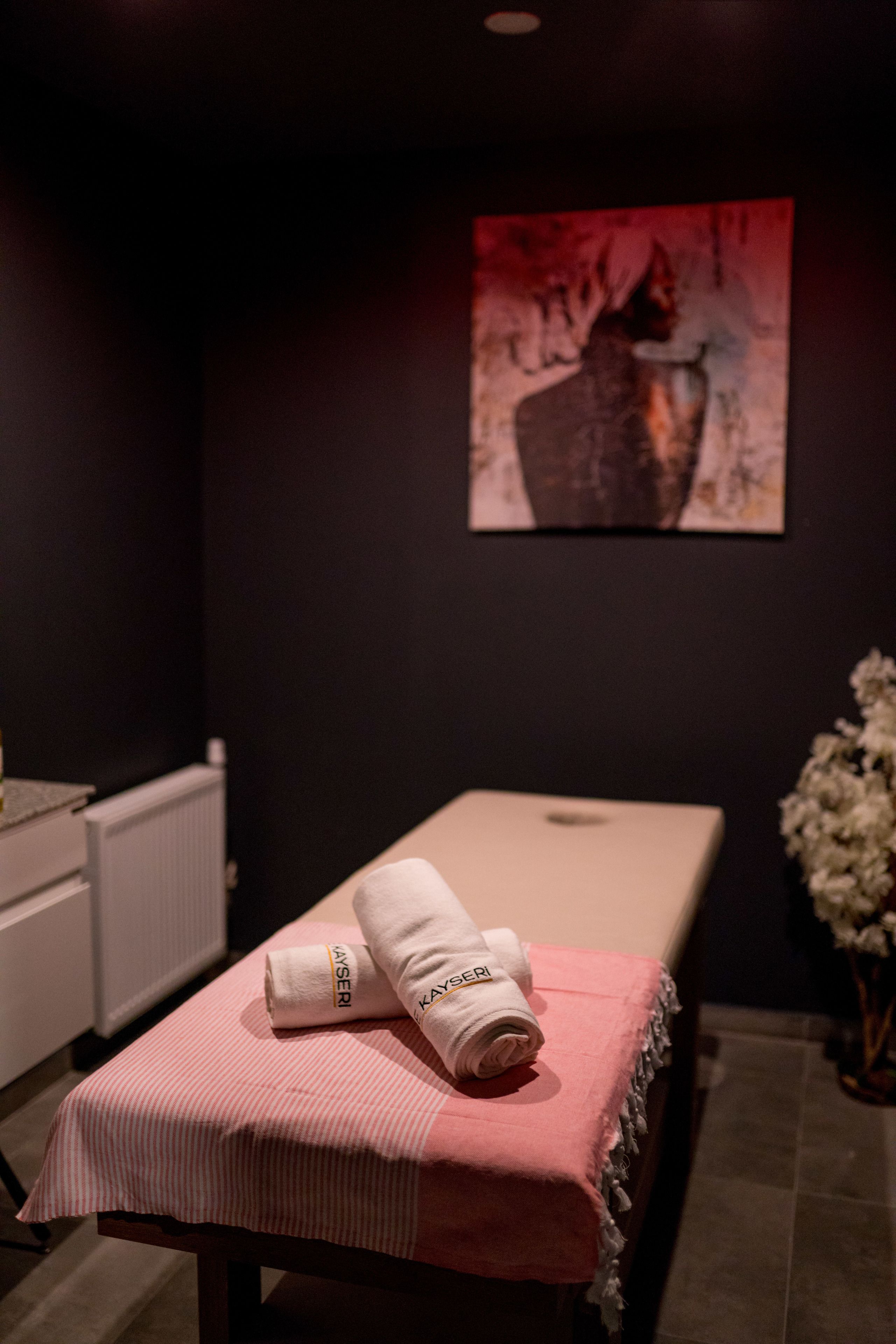 A room with a massage table covered by a pink towel, a white towel with a logo, a radiator, and a painting on the wall.