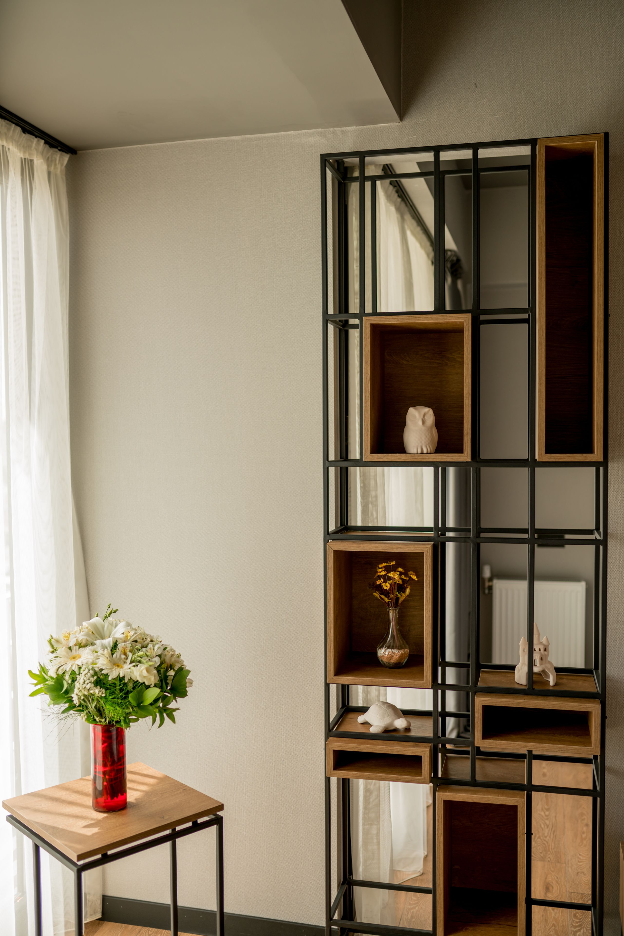 A room with a tall, black-framed shelving unit against a wall, a small wooden table with a red vase of flowers, and a white radiator.