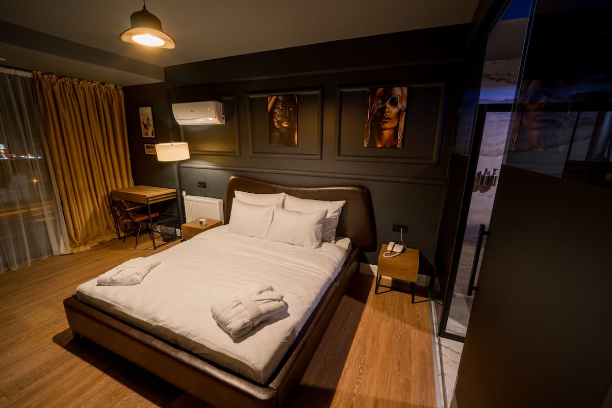 Ein Hotelzimmer mit einem großen Bett, einem Schreibtisch und einem Stuhl. Es gibt ein Fenster mit Vorhängen, eine Klimaanlage und ein Bild an der Wand.