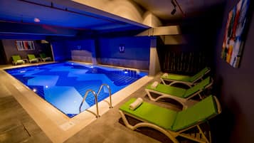 Ein Hallenbad mit blauem Boden, umgeben von Liegestühlen mit grünen Kissen.