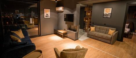 A modern living room with a sofa, armchair, and coffee table. There is a fireplace, a bookshelf, and artwork on the walls.