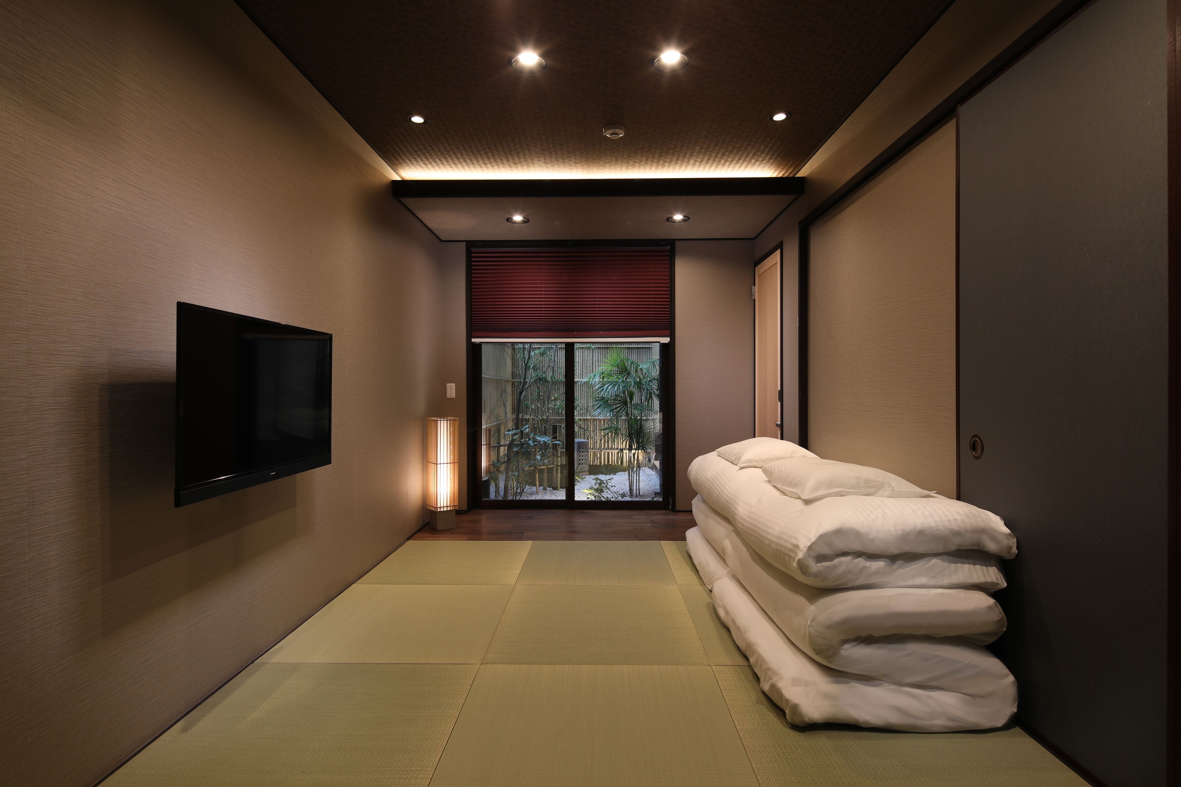 Habitación de hotel moderna con televisión de pantalla plana, esteras de tatami, una pila de toallas y una puerta corredera que da a un jardín.