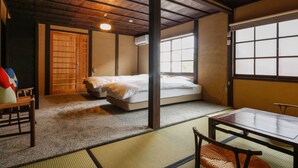 A traditional Japanese room with tatami flooring, a low wooden ceiling, a bed with white bedding, a table with chairs, and a sliding door. - Kariya Ryokan Q (Ako)