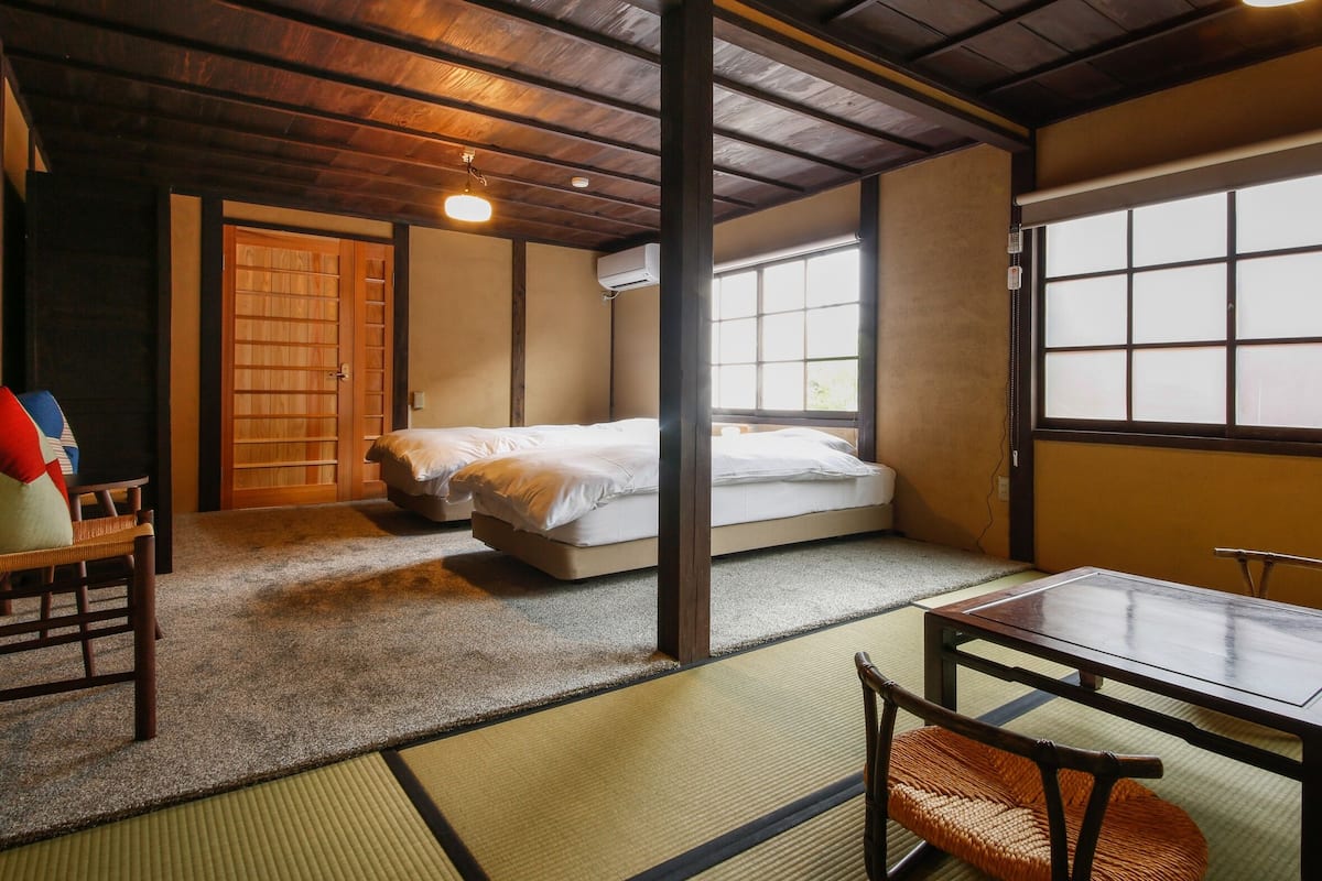 A traditional Japanese room with tatami flooring, a low wooden ceiling, a bed with white bedding, a table with chairs, and a sliding door.