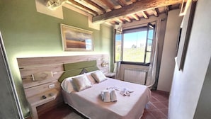 Un dormitorio con cabecera de madera, cama con cubrecama y almohadas blancas, mesita de noche y ventana con vistas a zonas verdes.