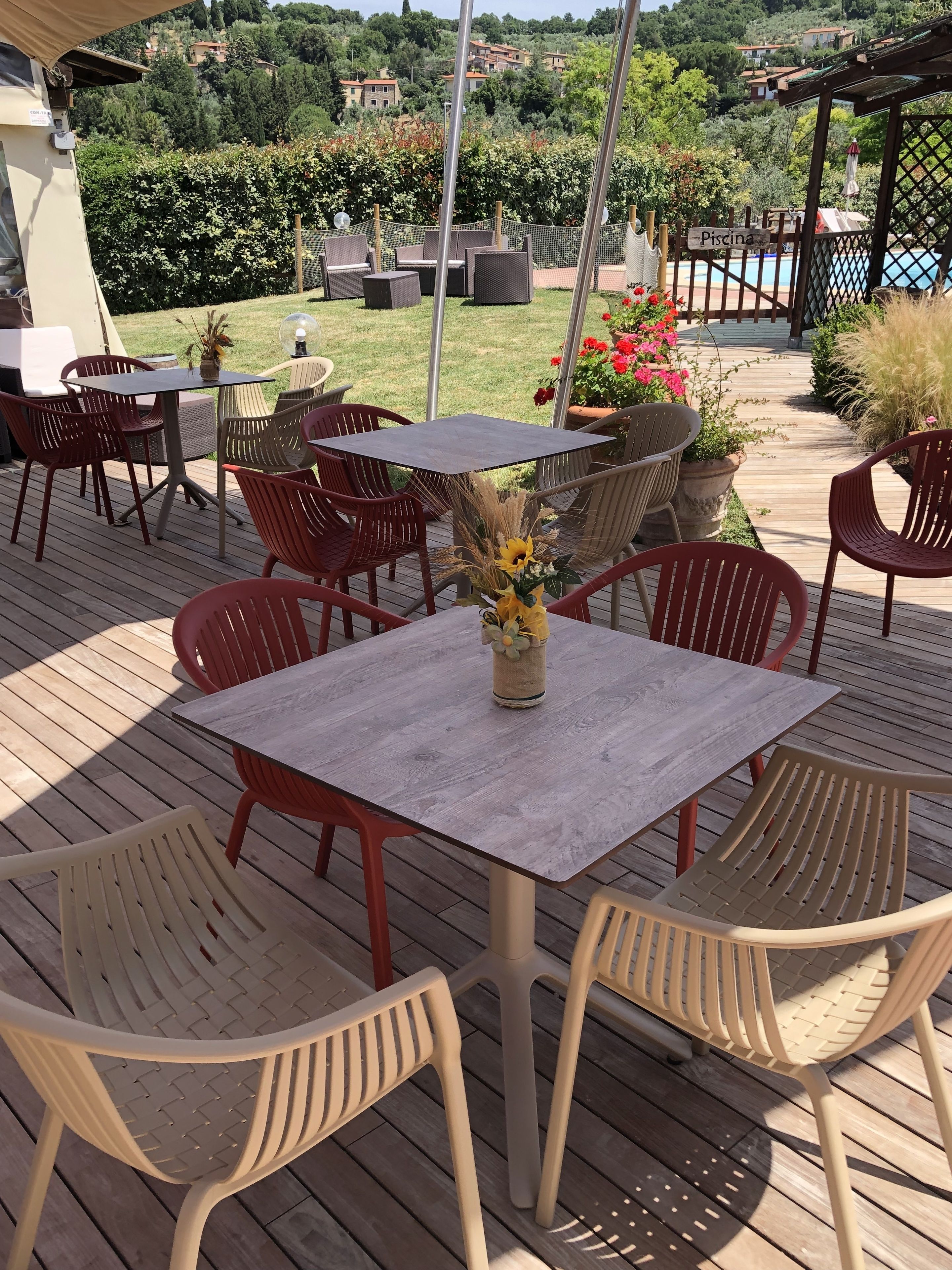 Een terras buiten met houten vloer, rode en beige stoelen en een tafel met een bloevas.