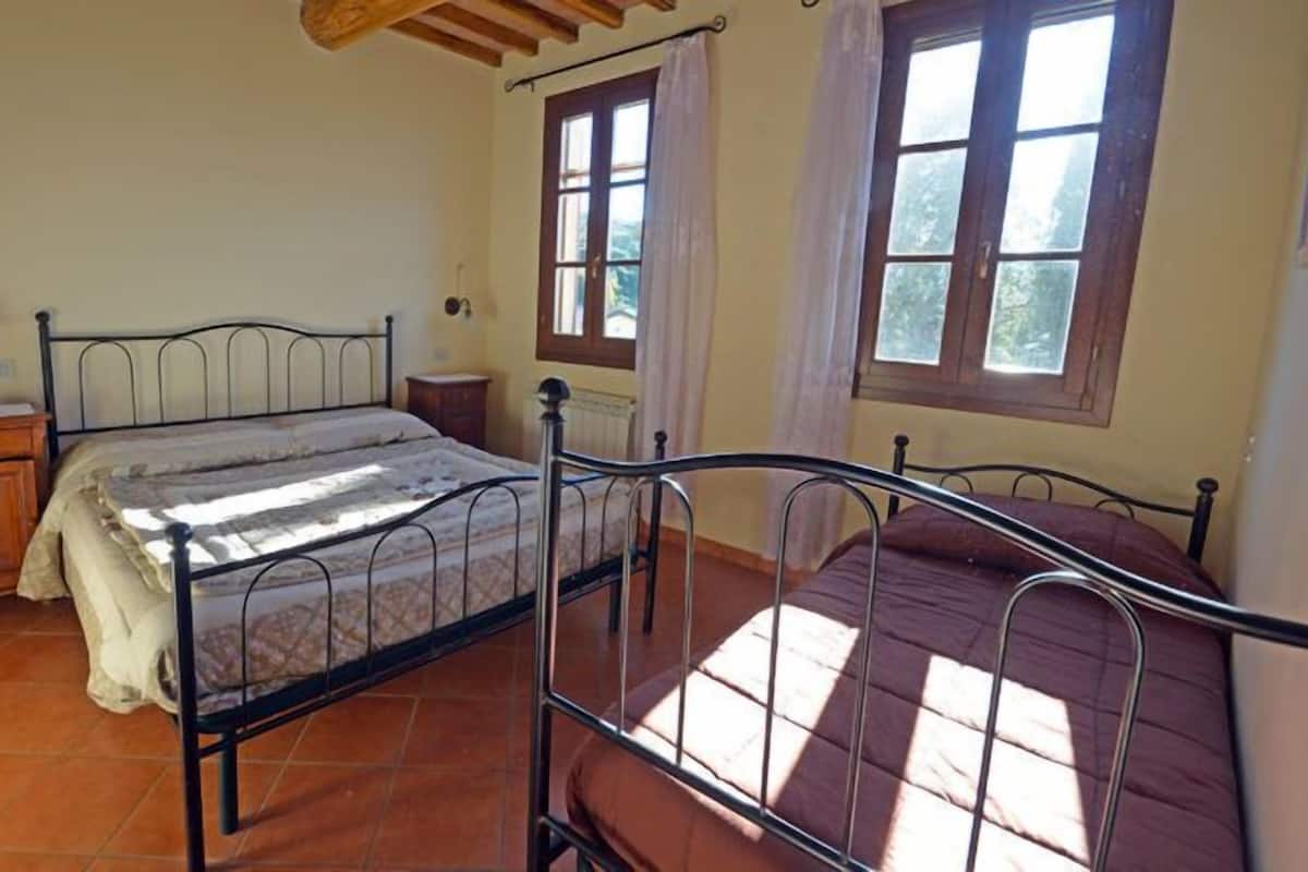 A bedroom with a metal bed frame, a wooden nightstand, a radiator, and two windows with curtains.