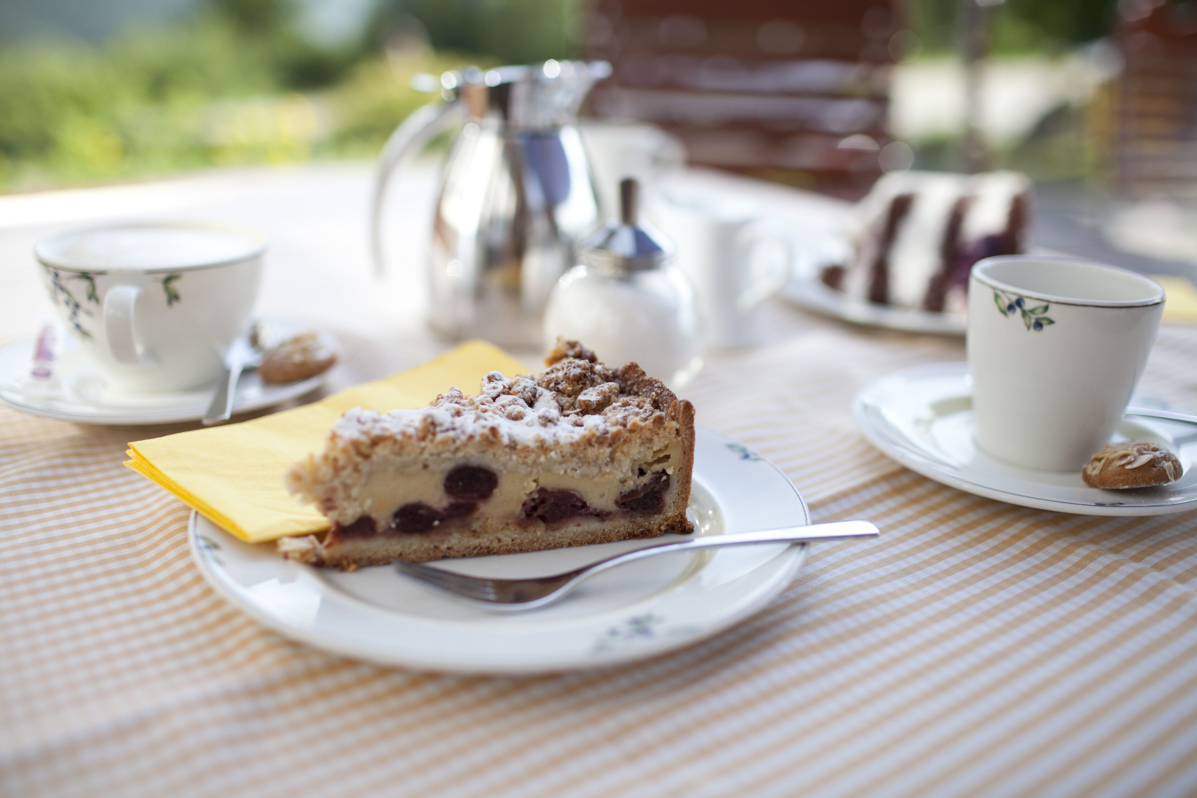 Ein Stück Kuchen auf einem Teller mit einem Löffel, einer Tasse und einer Kanne auf einem Tisch mit kariertem Tischtuch.