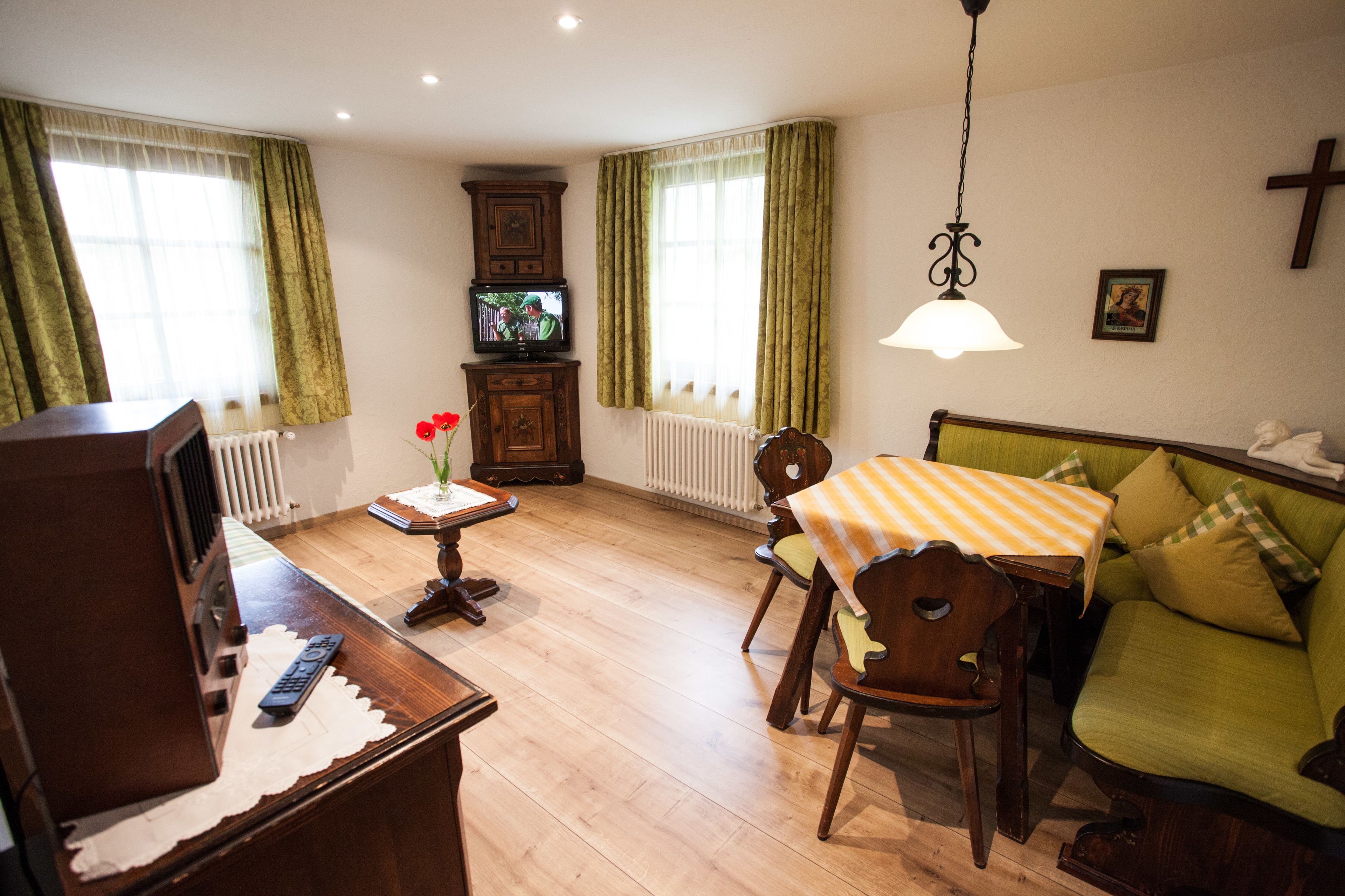 Ein Wohnzimmer mit Holzboden, einem grünen Sofa, einem Holztisch mit Streifenmuster, einer Holzkommode und einem Fernseher auf einem Standfuß.