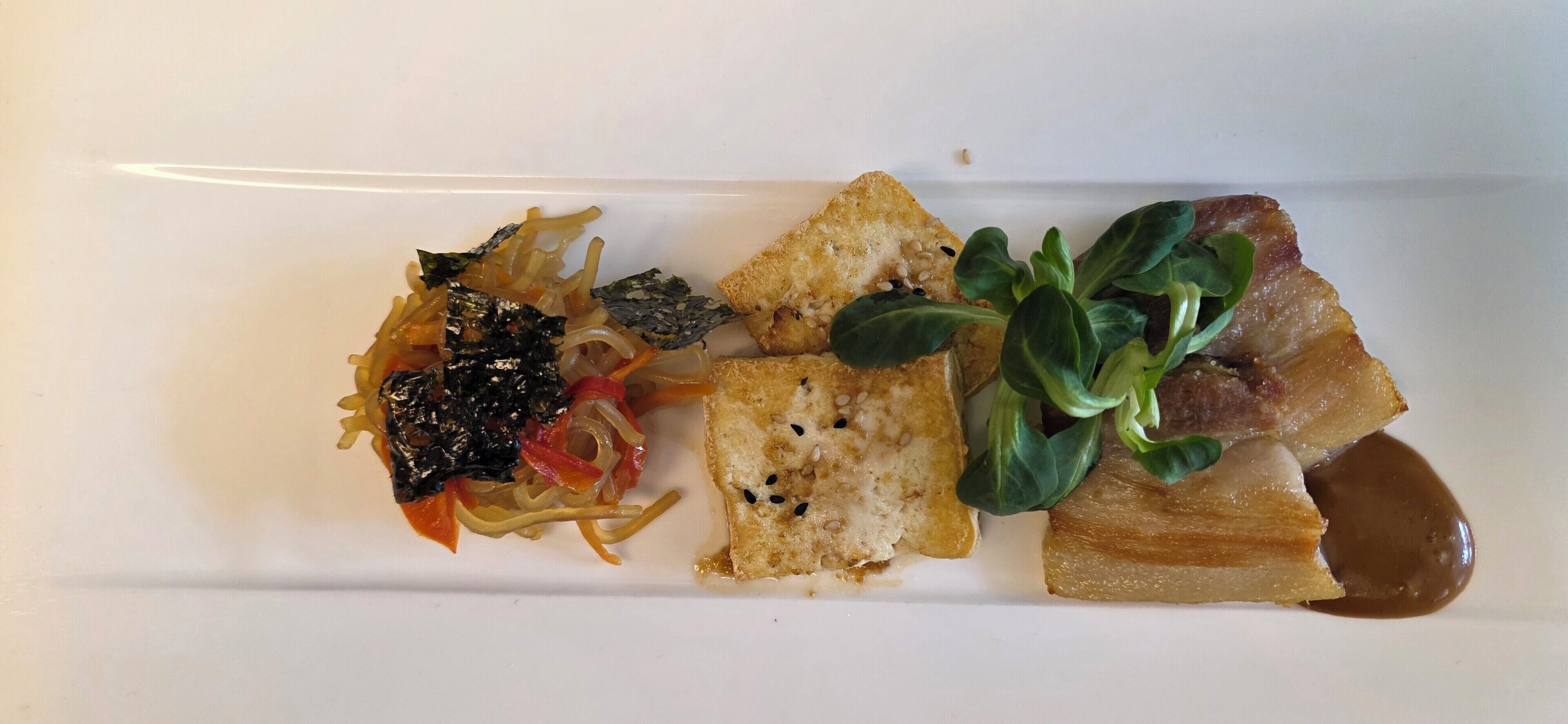 Un plato rectangular blanco con diferentes alimentos, como fideos, pan y un trozo de carne con salsa.