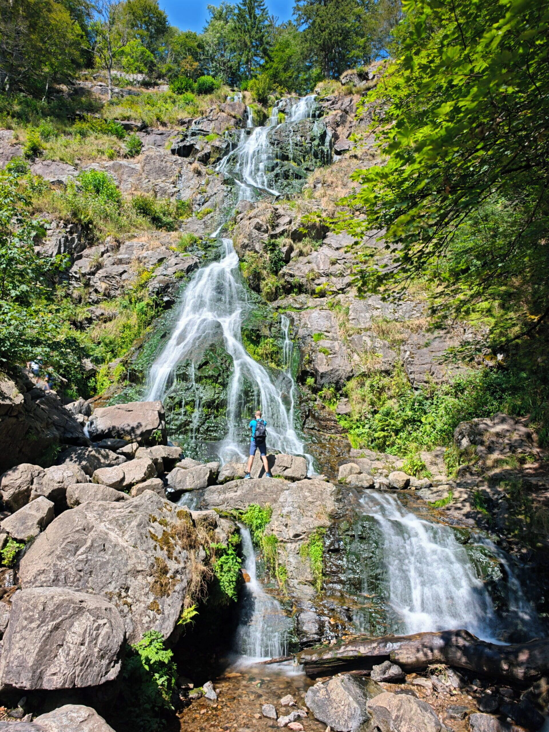 A waterfall cascading over rocks in a forested area with a person standing on a rock.