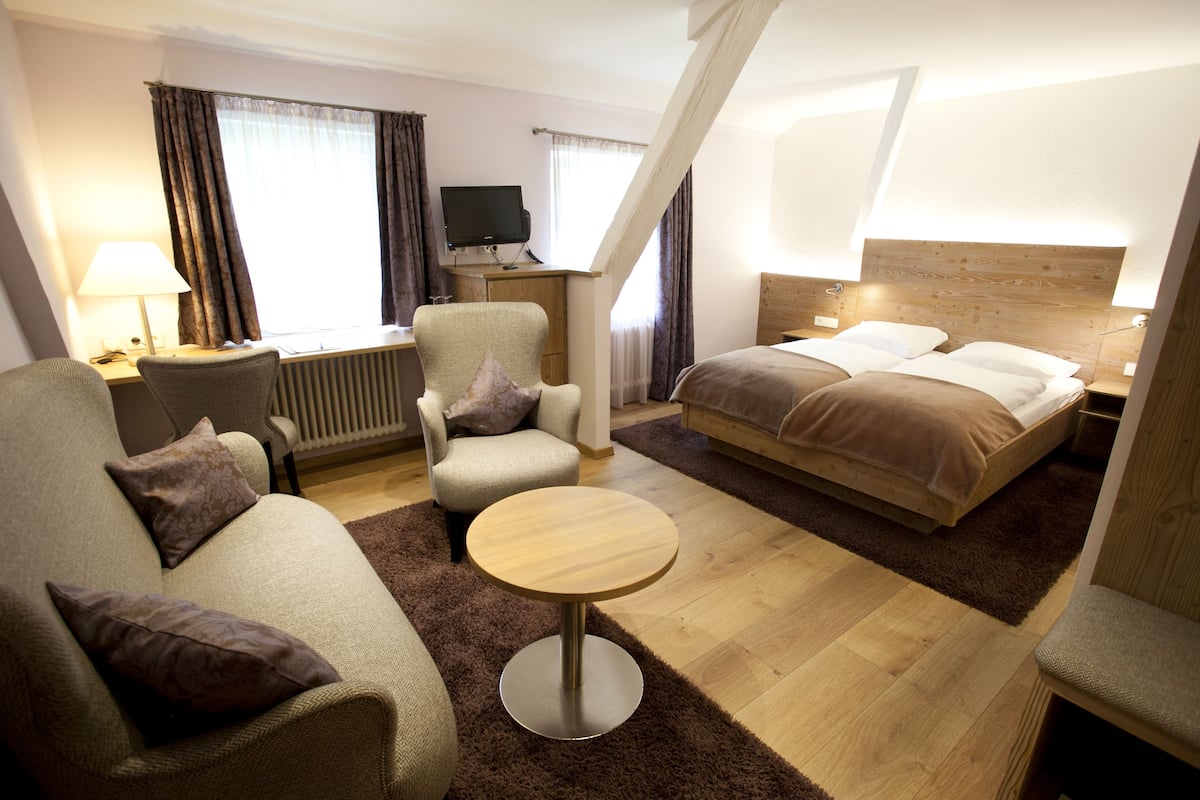 A hotel room with a bed, a sofa, a chair, a small table, a desk with a lamp, and a TV.