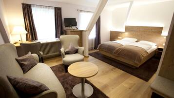 Una habitación de hotel con cama, sofá, sillón, una mesita, escritorio con lámpara y televisor.
