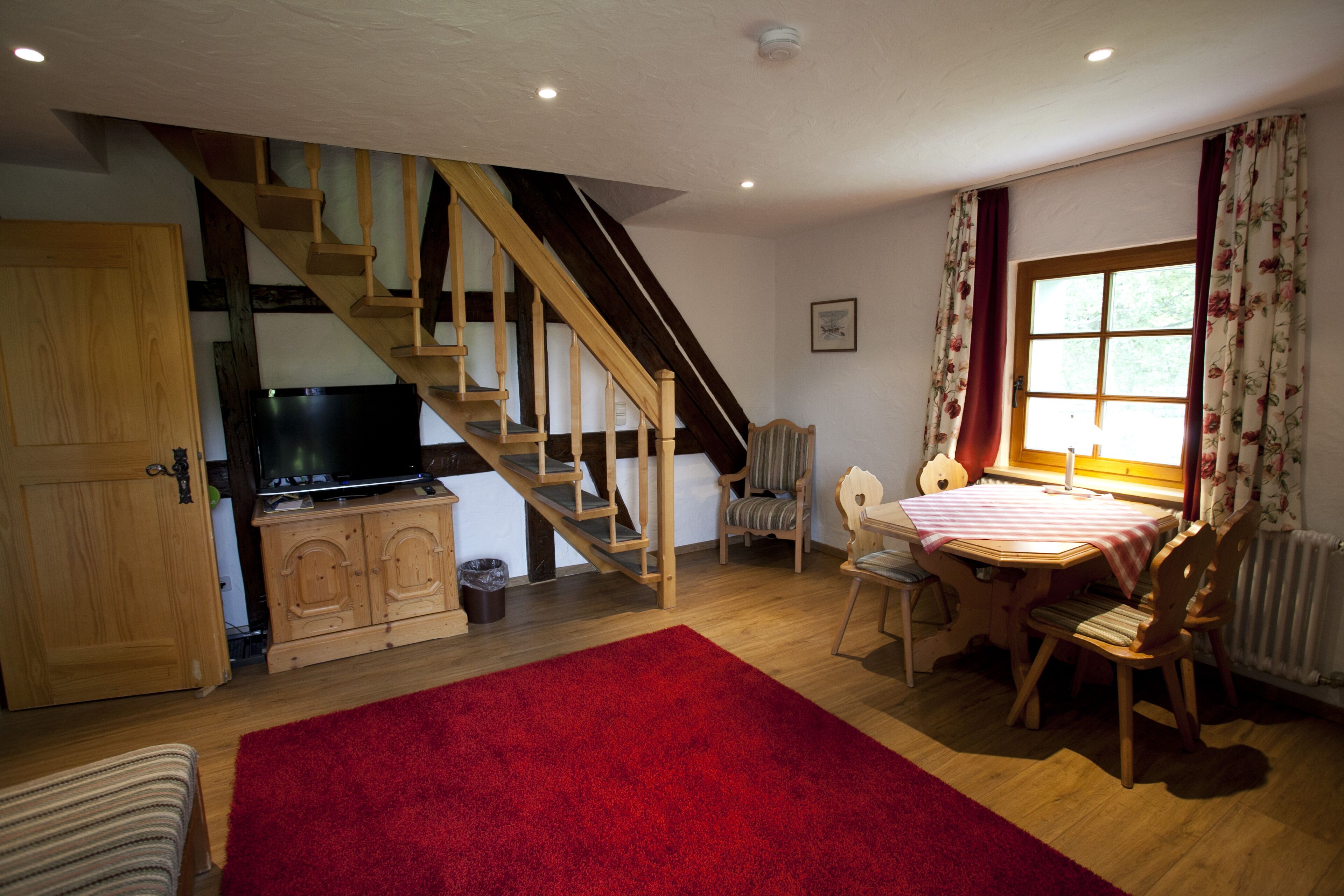 A cozy living room with a wooden staircase, a television, a dining table with chairs, and a red rug.
