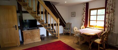 A cozy living room with a wooden staircase, a television, a dining table with chairs, and a red rug.