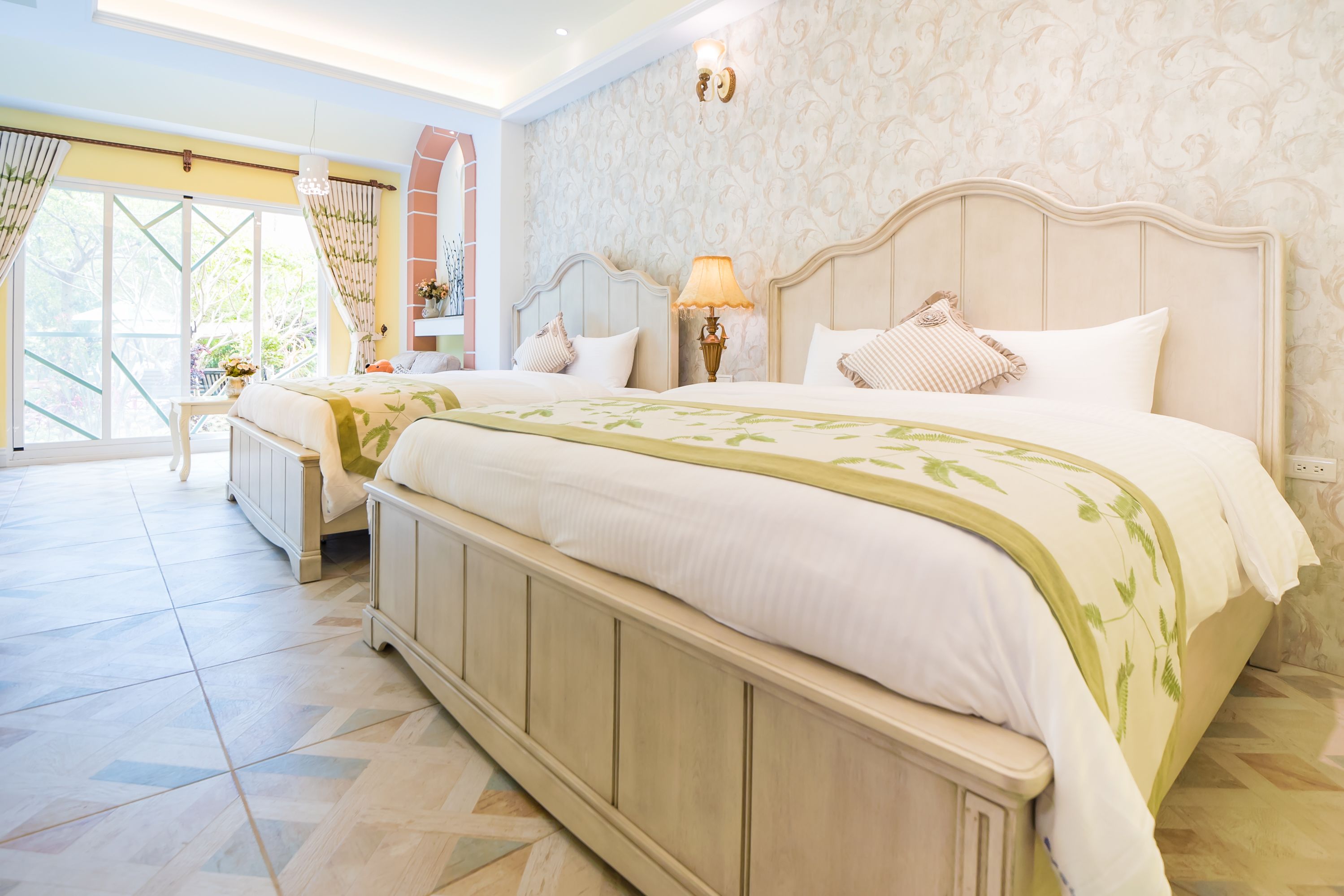 A spacious bedroom with a large bed, a nightstand, a lamp, and a view of the outdoors through a large window.