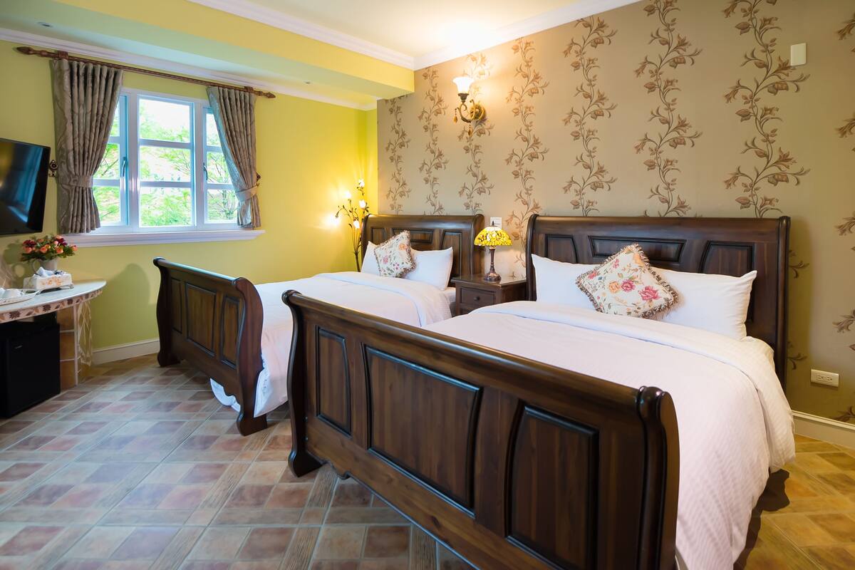 A double bed with a wooden headboard, a bedside table with a lamp, and a floral-patterned wall.