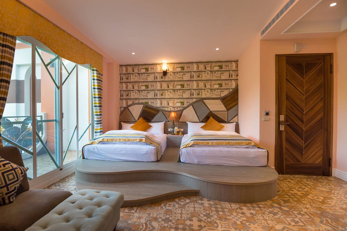 A hotel room with two beds, a large window, a wall-mounted bookshelf, and a wooden door.