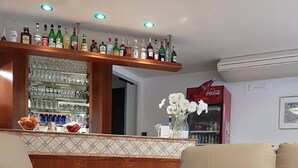 Eine Bar mit verschiedenen Spirituosenflaschen und Gläsern, einem Coca-Cola-Kühlschrank sowie einer Sitzecke mit zwei beigen Sofas und einem runden Tisch.