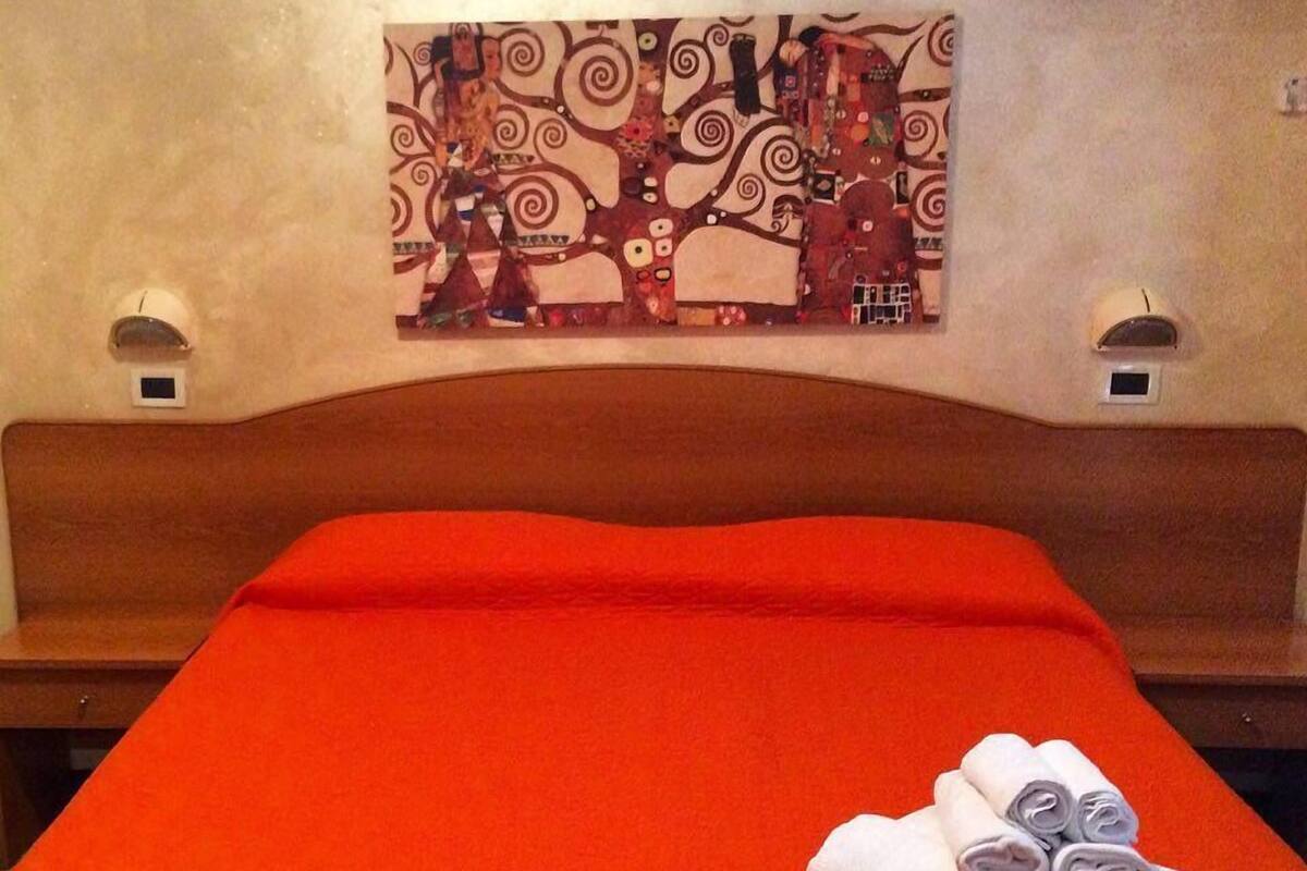 A hotel room with a red bedspread, a headboard, a wall-mounted artwork, and two wall sconces.