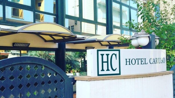 Ein Hotel Eingang mit einem Schild, auf dem 'HOTEL CARUSO' steht, und einer großen Sonnenschirm-Struktur im Freien.