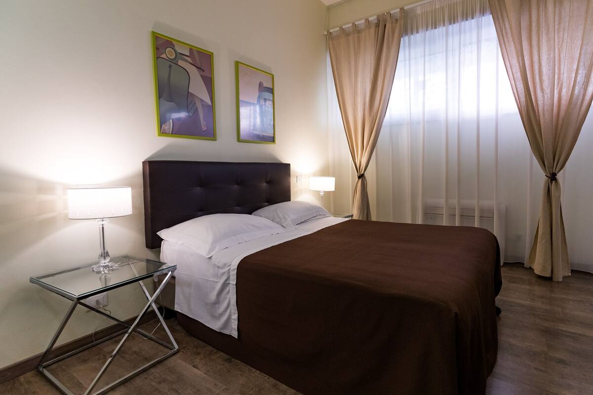 A hotel room with a bed, a glass side table, a lamp, and two framed pictures on the wall.