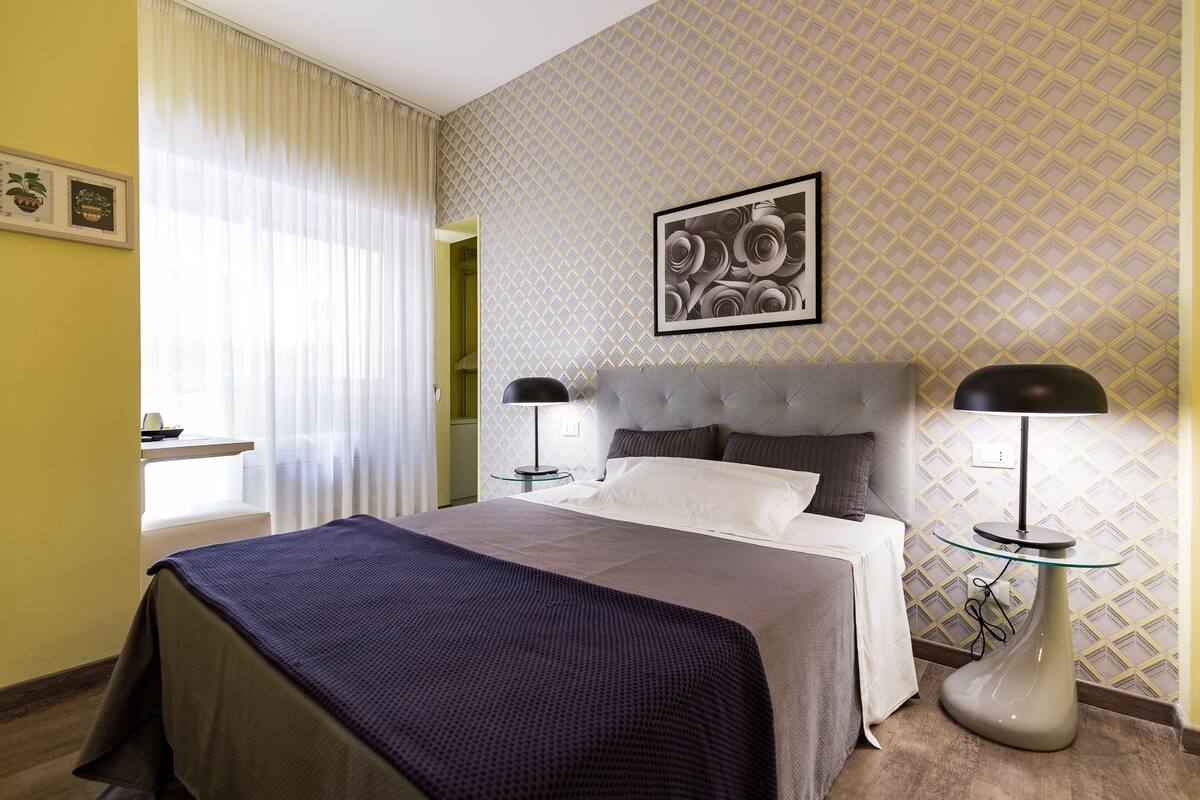 A hotel room with a bed, two bedside tables with lamps, a framed picture on the wall, and a window with sheer curtains.