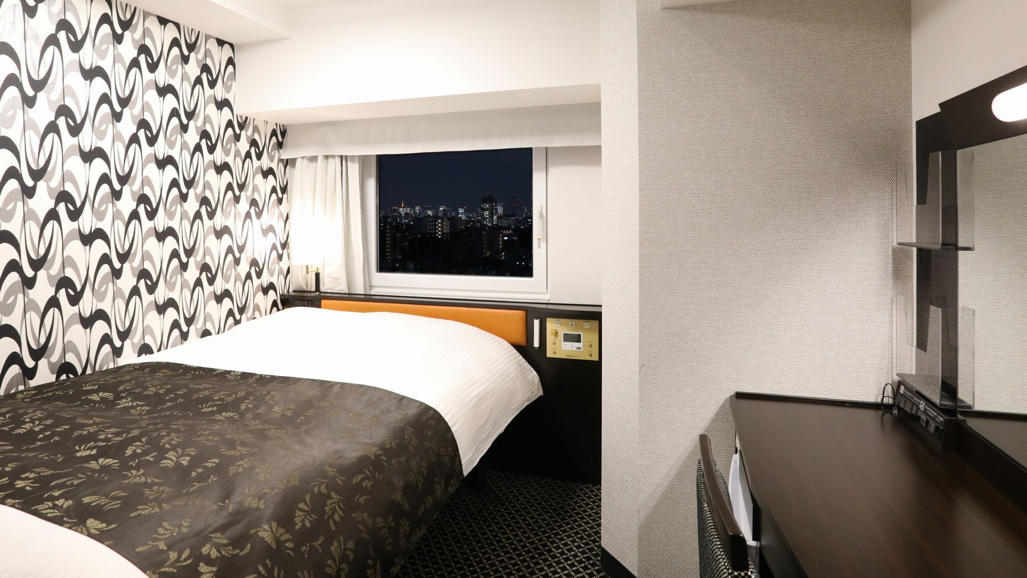 A hotel room with a bed, a window with a city view, a bedside table, and a mirror.